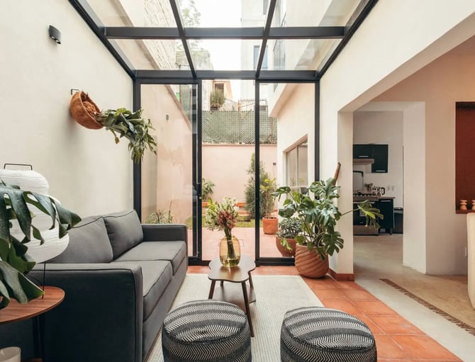 Glass-ceiling living room with terracotta tile floors, monstera plants, a grey sofa, and a view through to a courtyard garden